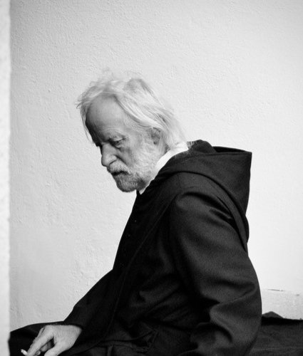 Klaus Huhle © Kerim Waller, Portraitfoto des Schauspielers Klaus Huhle, er sitzt mit Zigarette vor einer Wand, trägt einen schwarzen Überwurf und blickt diagonal zu Boden. Er trägt einen weißen Bart, seine Haare reichen bis in den Nacken